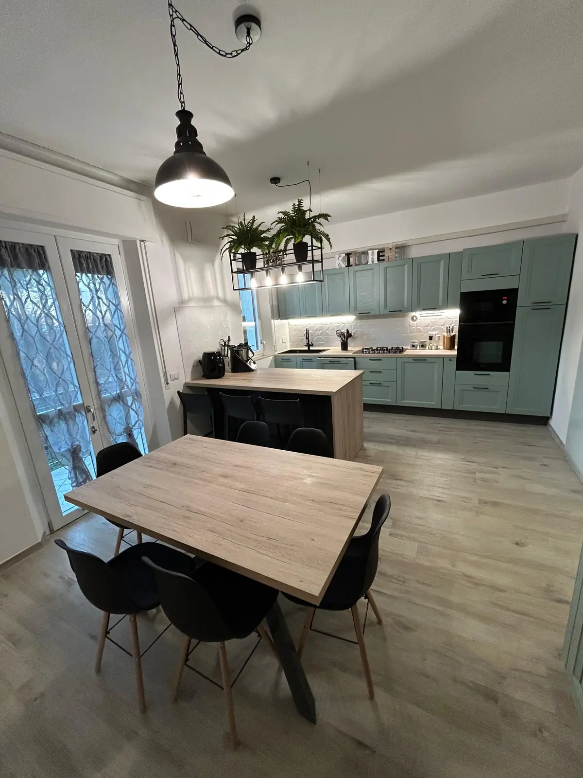 Cucina verde salvia con isola, colonna forno e tavolo da pranzo in un open space